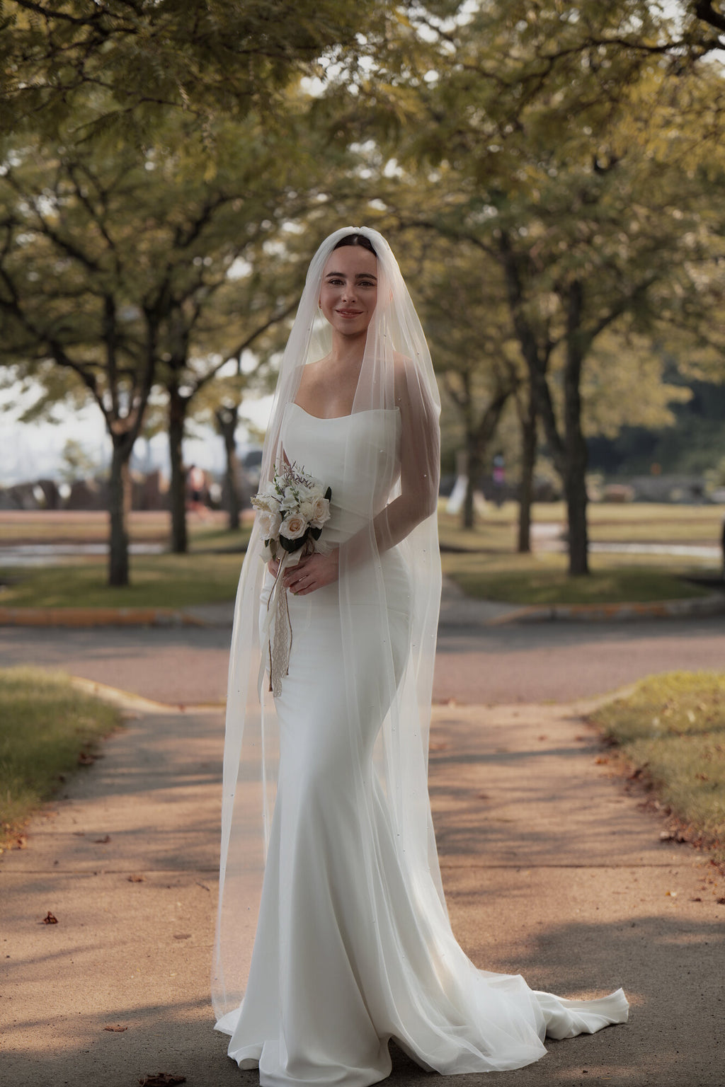 Bride outdoors holding a bouquet, wearing The Soft Focus scattered pearl cathedral veil over a white column gown
