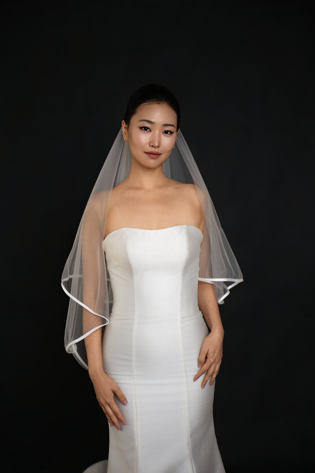 Front view of The Clean Line two tier fingertip veil with a narrow satin ribbon edge, blusher worn back, over a strapless white gown