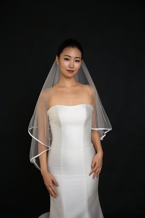 Front view of The Clean Line two tier fingertip veil with a narrow satin ribbon edge, blusher worn back, over a strapless white gown