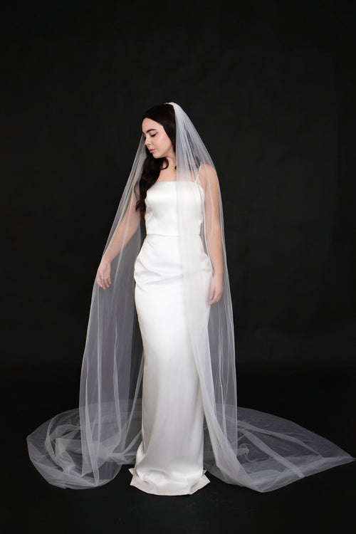 Full length front view of The Barely There cathedral veil in white tulle pooling on the floor beside a strapless satin column gown, on a black backdrop