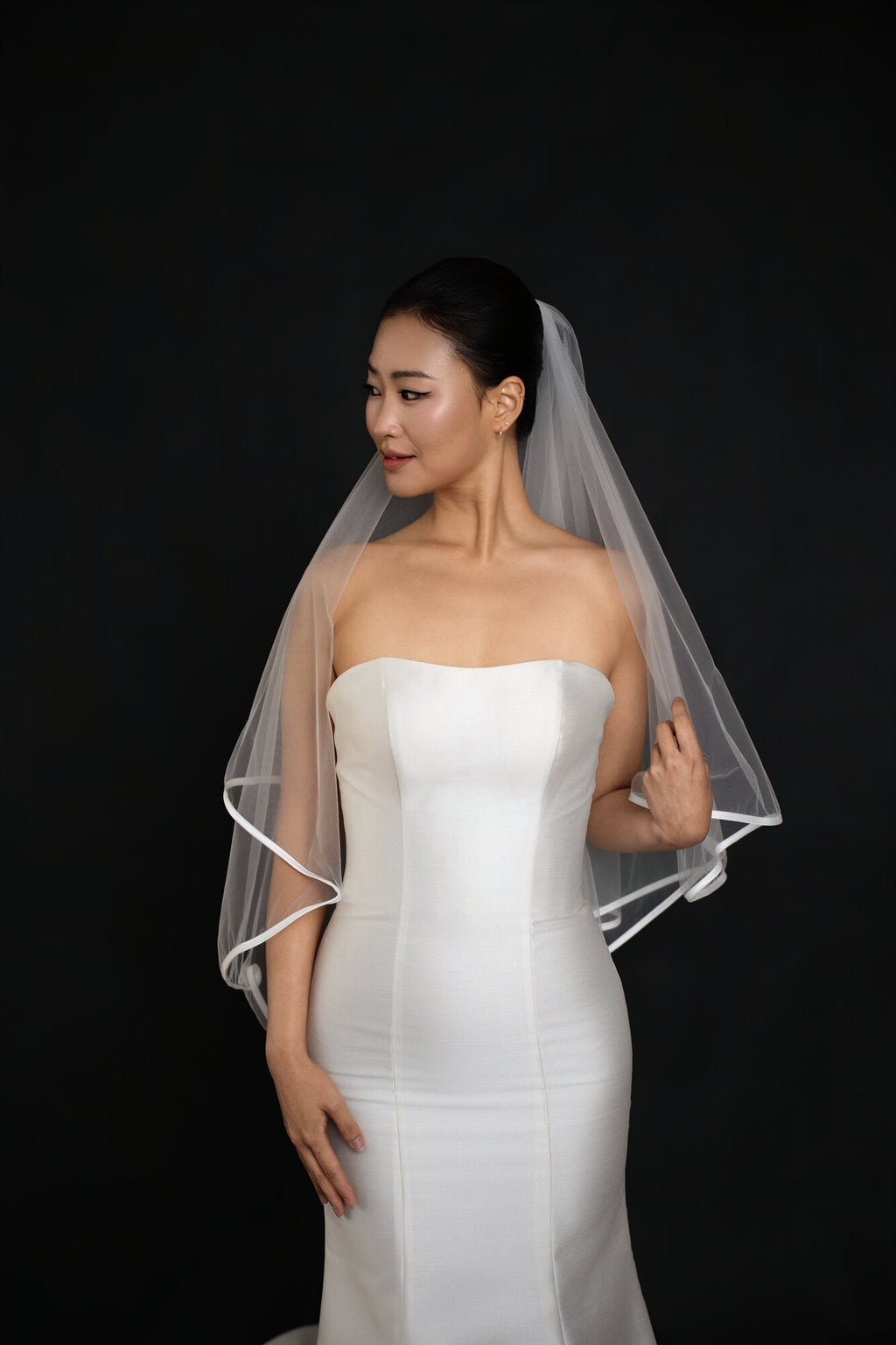 Bride adjusting The Clean Line satin edge veil, three quarter view showing both tiers and the ribbon border