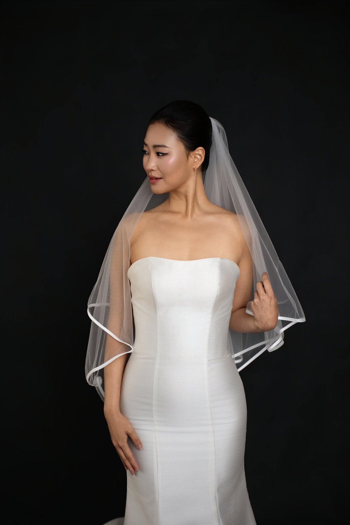 Bride adjusting The Clean Line satin edge veil, three quarter view showing both tiers and the ribbon border