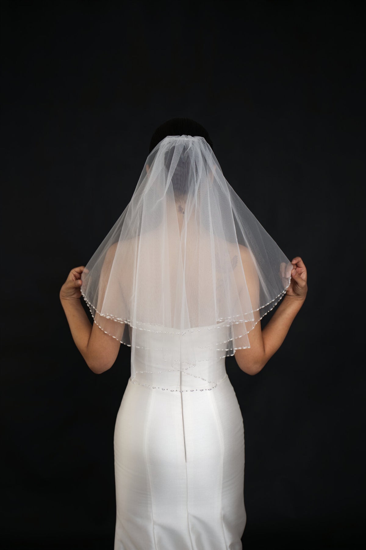 Back view of The Second Look veil with the bride lifting the blusher layer, pearl edge catching light on a black backdrop