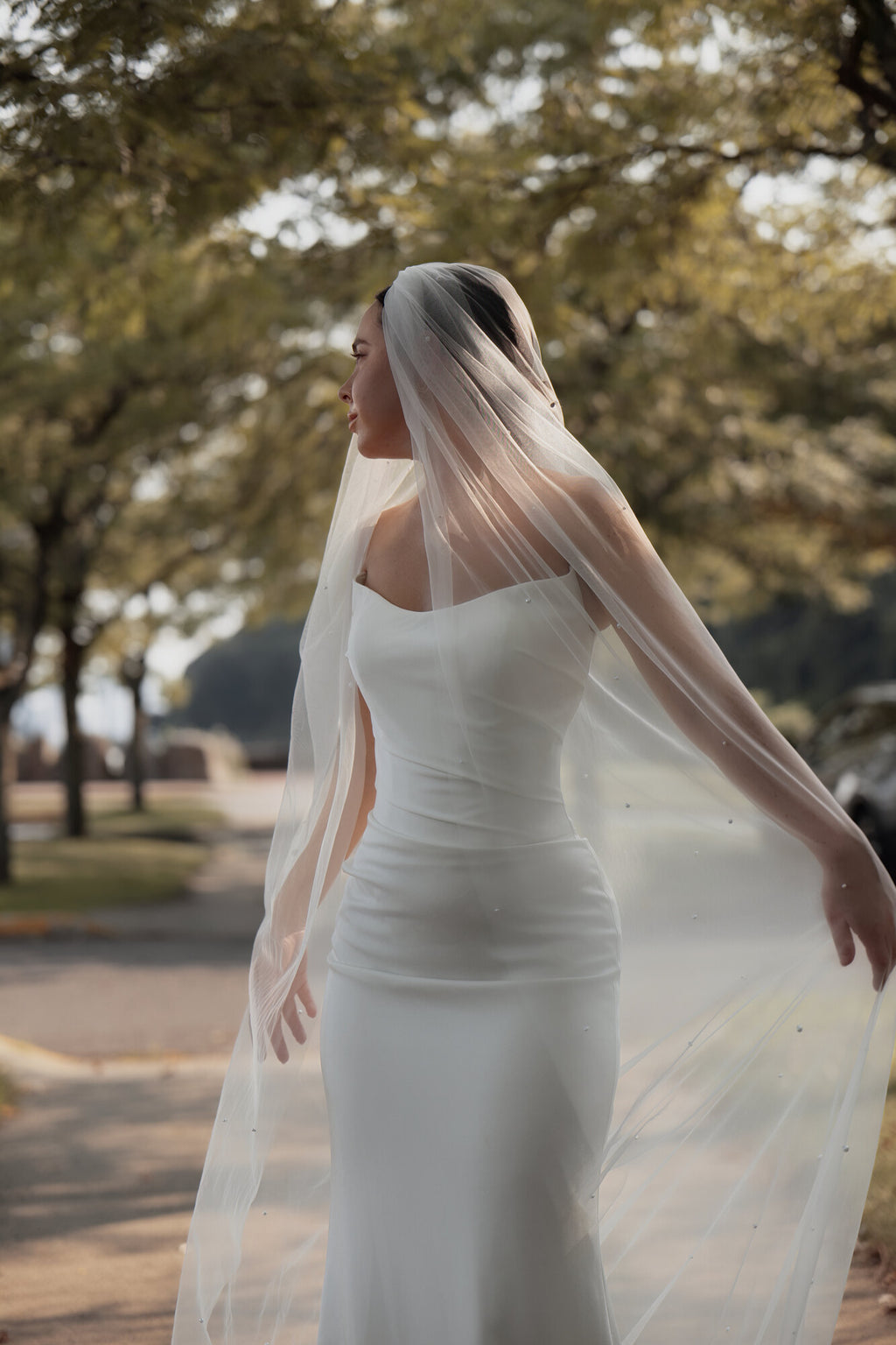 The Soft Focus pearl cathedral veil photographed outdoors in daylight, tulle lifting behind a white satin gown