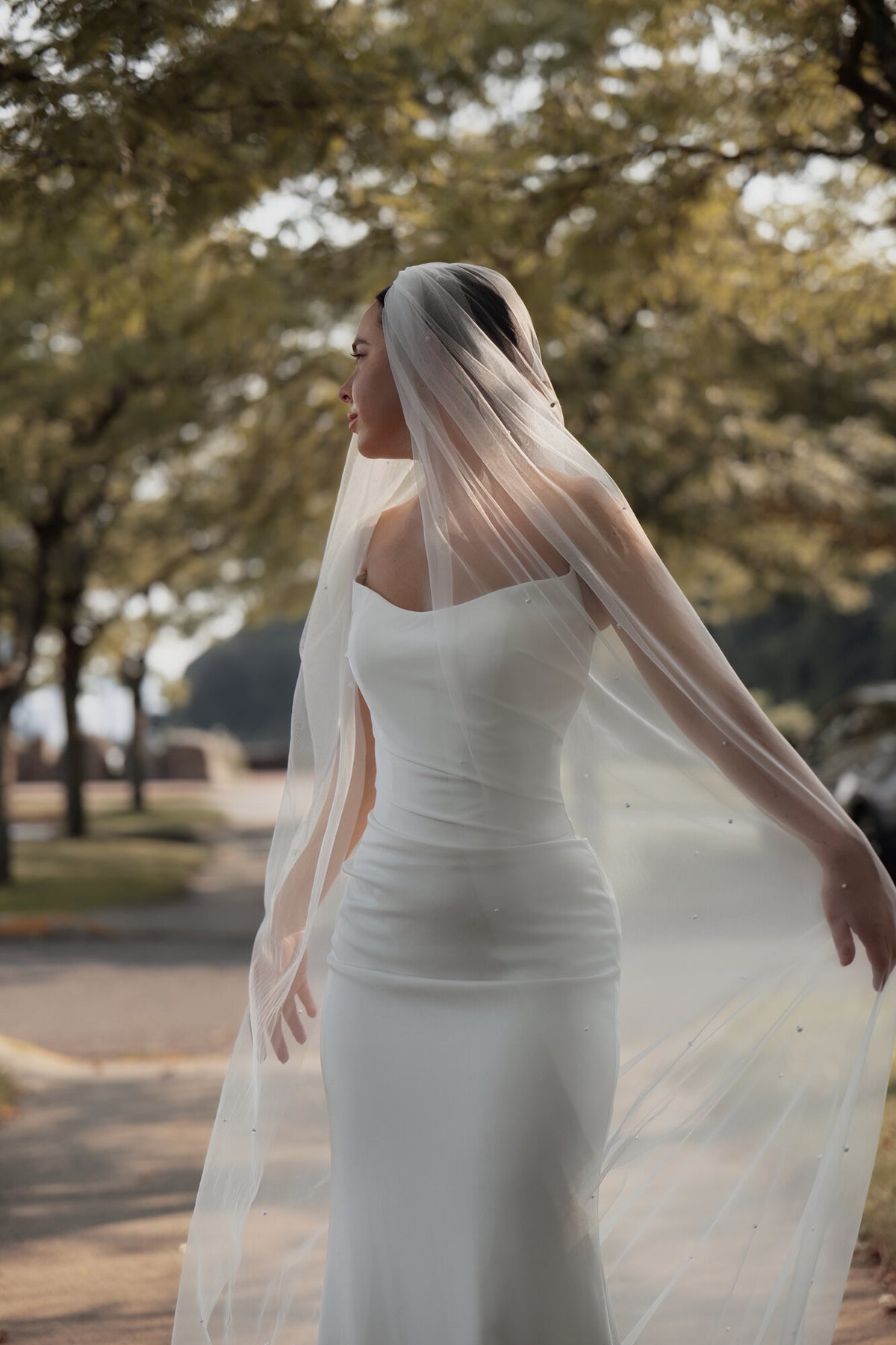 The Soft Focus pearl cathedral veil photographed outdoors in daylight, tulle lifting behind a white satin gown