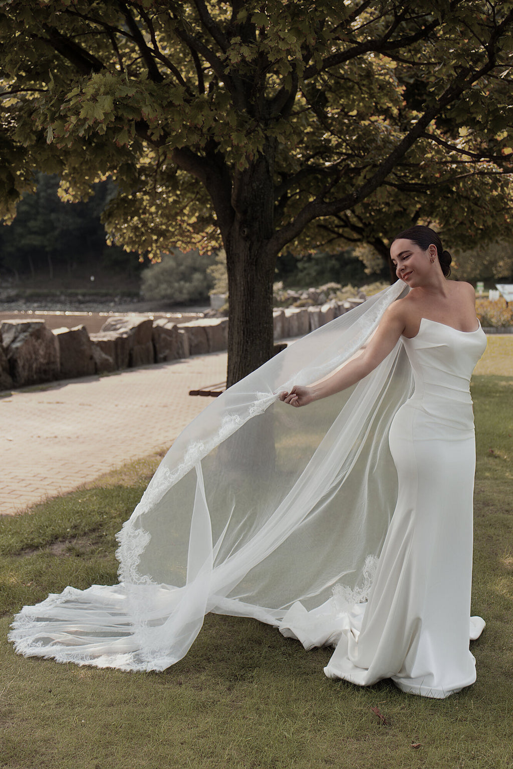 Bride outdoors holding The Close Up mantilla veil out to one side, lace border trailing across the grass
