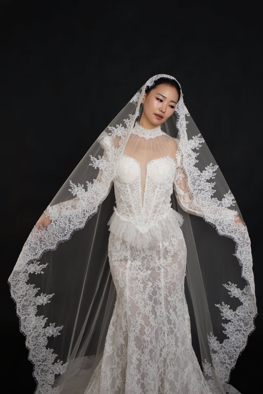 A woman wearing a white lace bridal gown with a long sheer veil featuring floral lace trim, posed against a solid black background.
