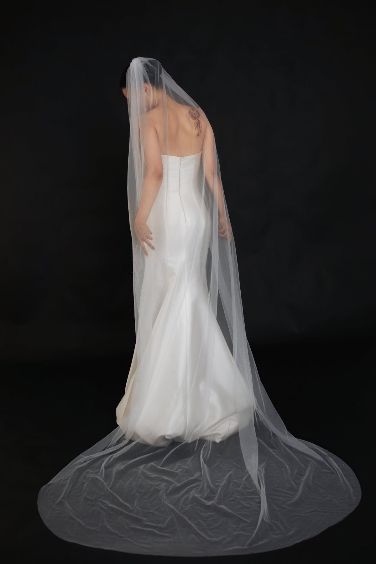 A woman wearing a strapless white wedding gown and a long sheer bridal veil, photographed from the back against a solid black studio background.