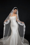 A woman wearing a strapless white mermaid-style wedding gown and a long sheer white veil with lace trim, posed against a solid black studio background.