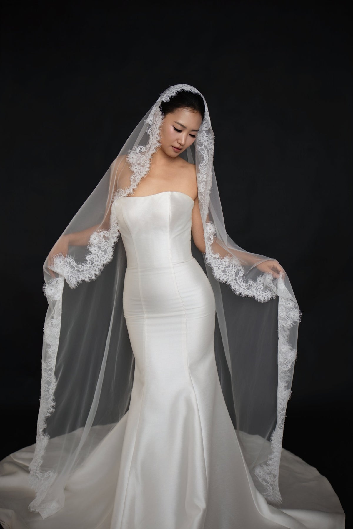 A woman wearing a strapless white mermaid-style wedding gown and a long sheer white veil with lace trim, posed against a solid black studio background.