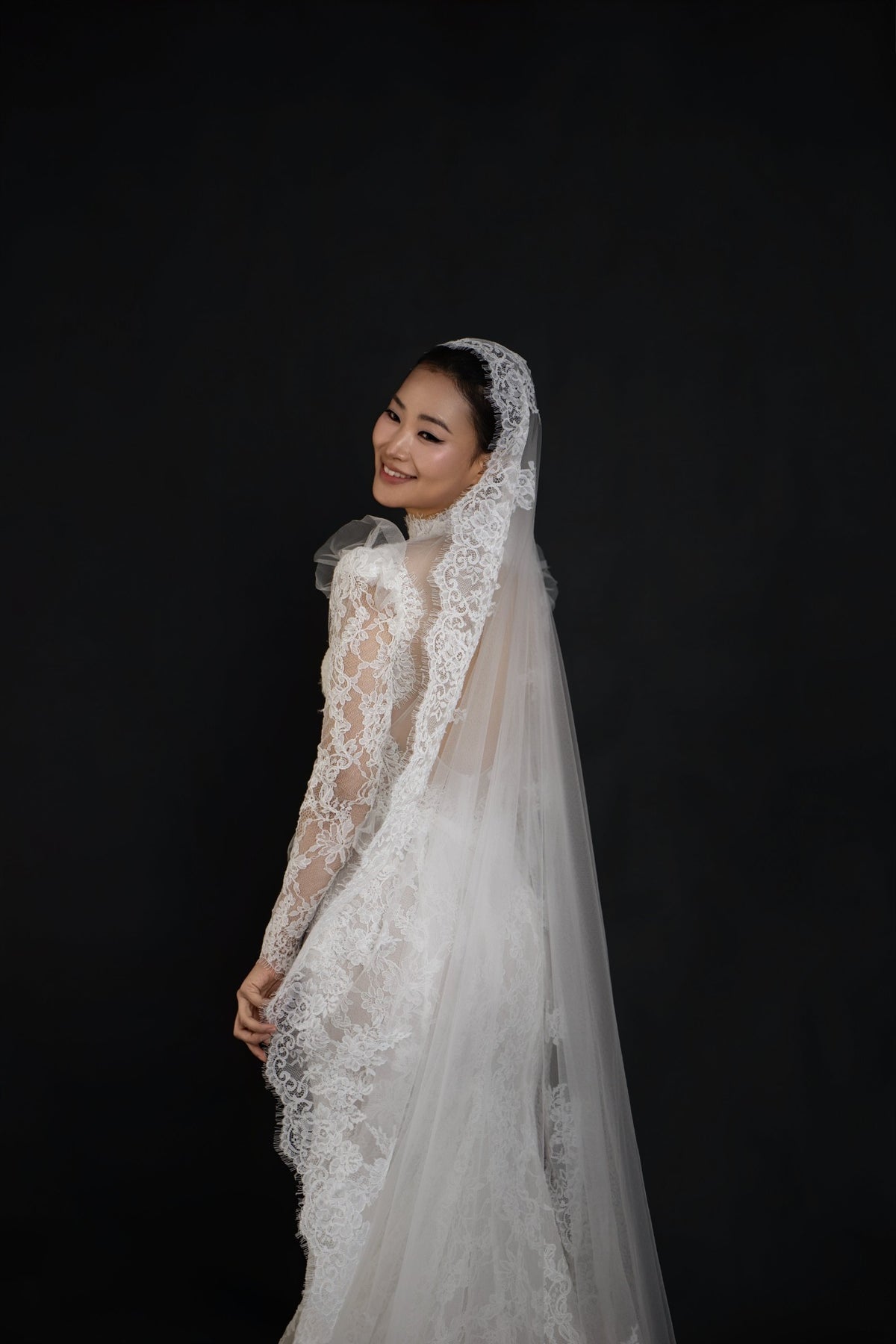 A woman wearing a white lace bridal gown with long sheer sleeves and a matching veil, posed against a solid black background.