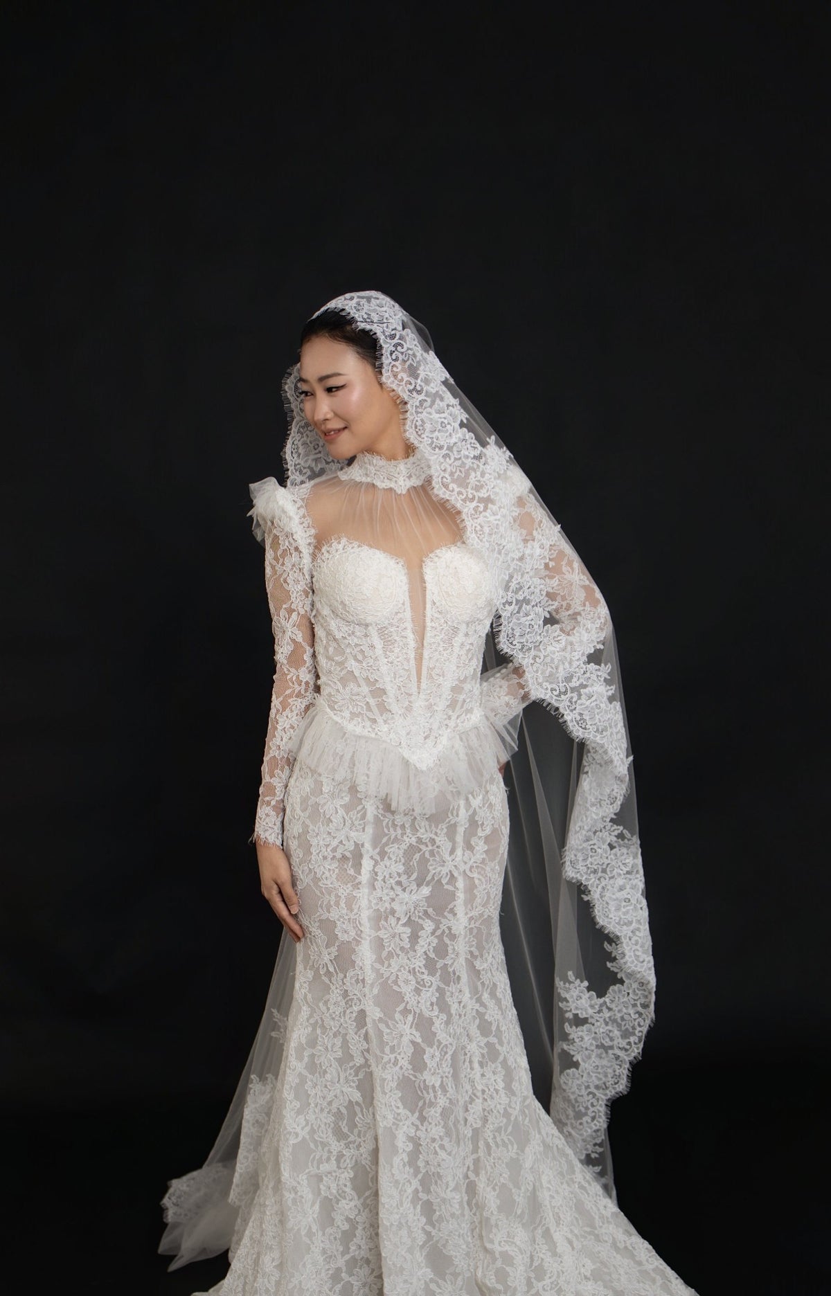 A woman modeling a white lace wedding gown with a sheer lace veil against a solid black studio background.