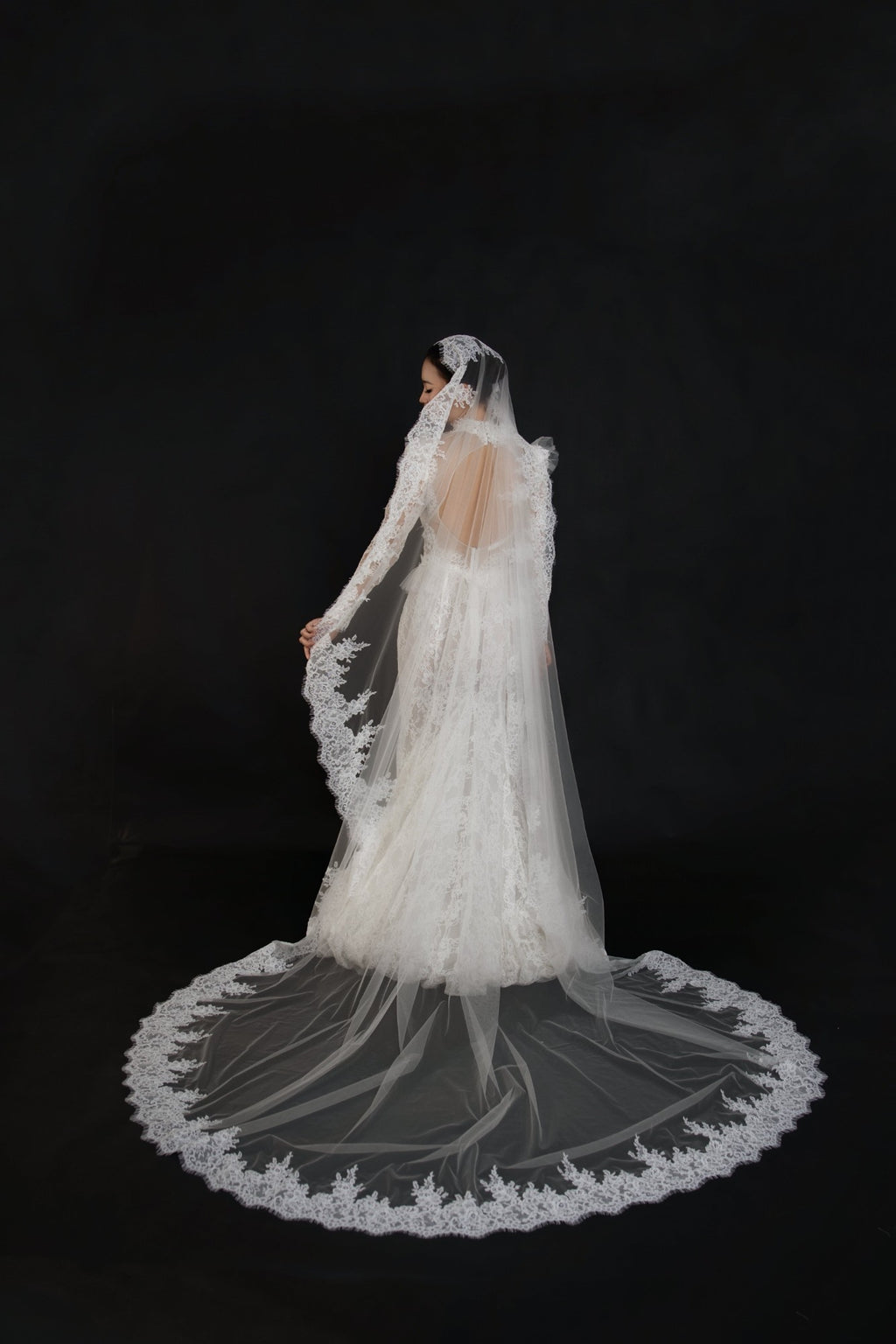 A bride wearing a long-sleeved lace wedding gown and matching sheer veil with a large circular train, photographed from the back against a solid black background.
