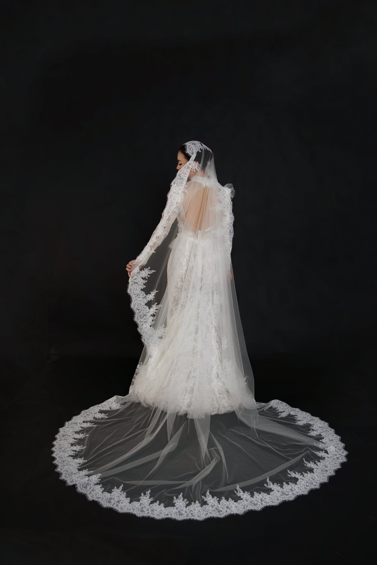 A bride wearing a long-sleeved lace wedding gown and matching sheer veil with a large circular train, photographed from the back against a solid black background.