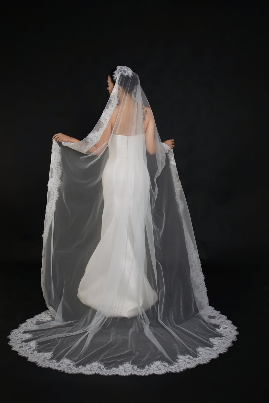 A bride shown from the back wearing a long sheer white tulle bridal veil with floral lace trim, held out to show its full length and shape against a black studio background.