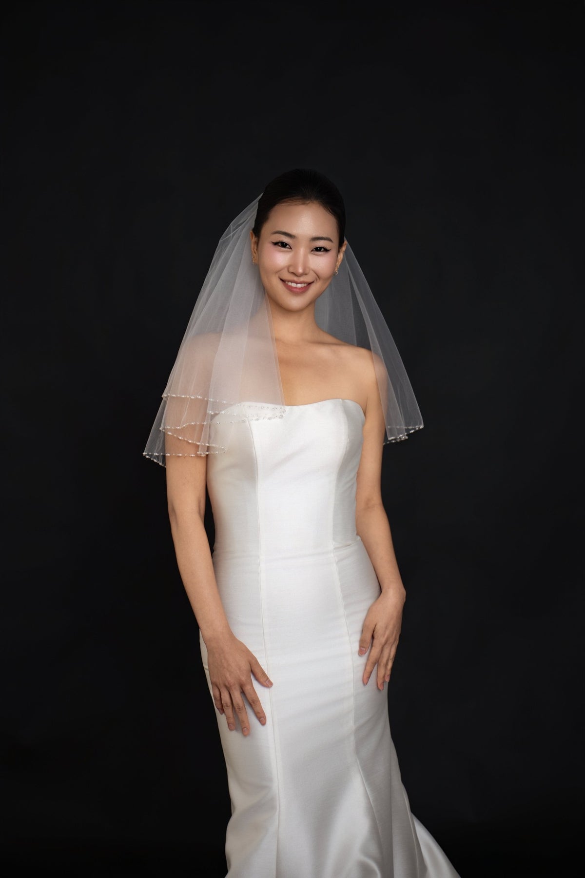 A woman wearing a white strapless wedding gown and a sheer white veil, standing against a solid black background.